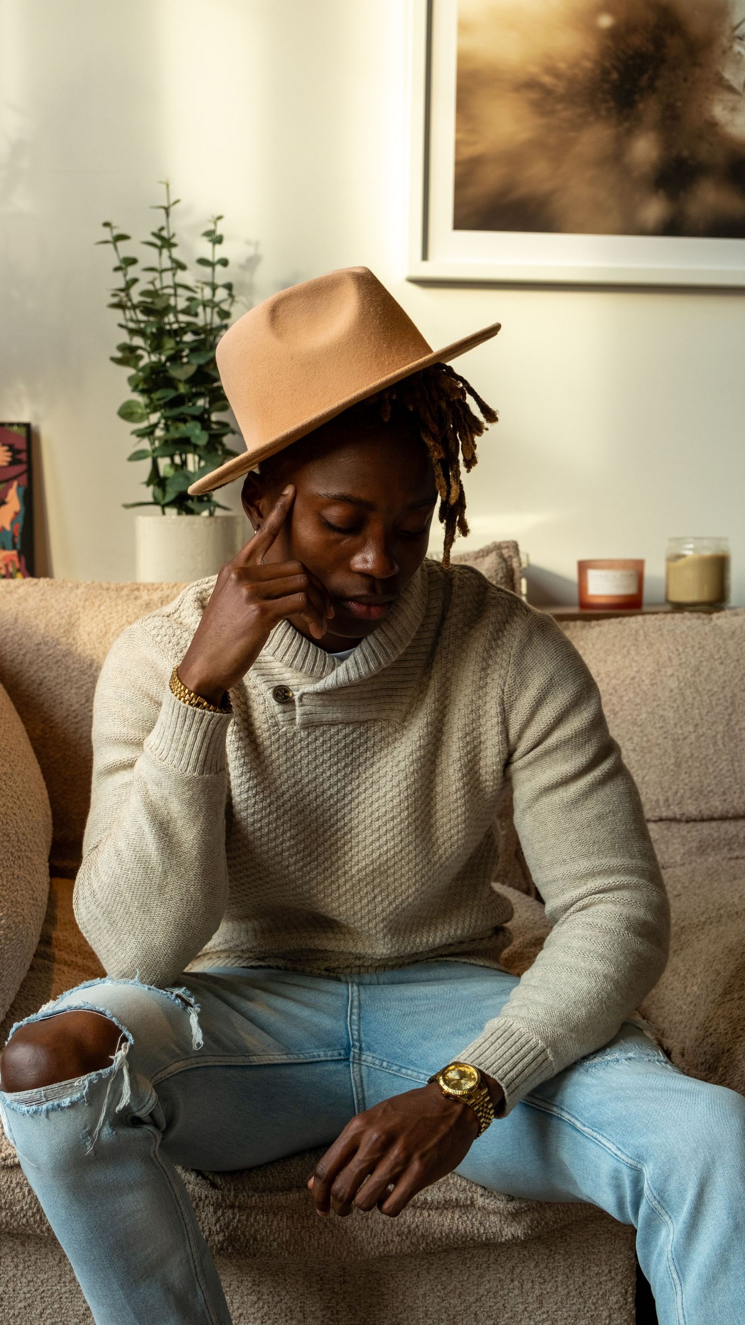 Stylish man in beige sweater and ripped jeans wearing a tan fedora hat and gold watch.