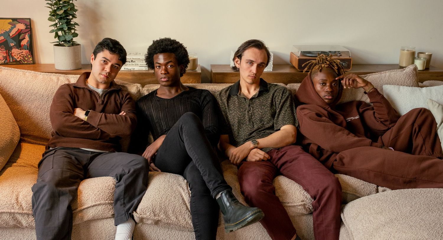 Four young men sitting casually on a couch together in a bright living room setting.
