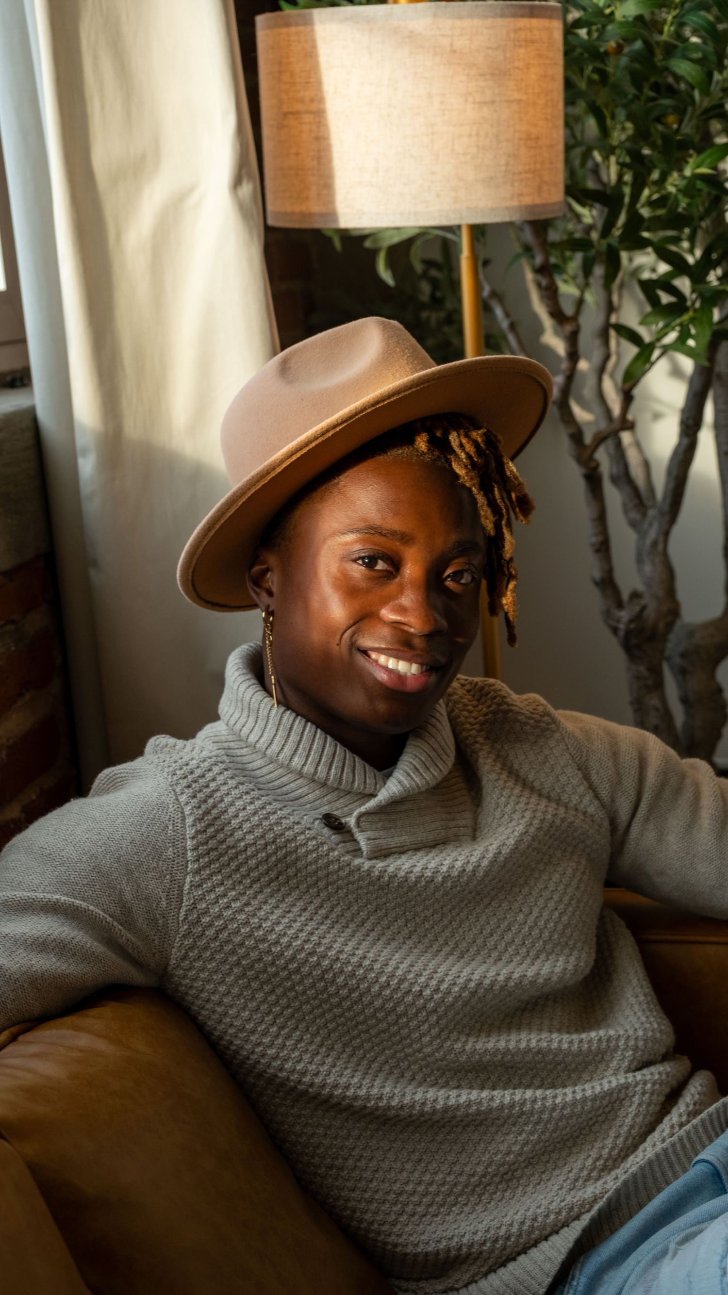 Smiling Black man wearing tan wide-brim hat and gray turtleneck sweater seated on leather couch indoors.