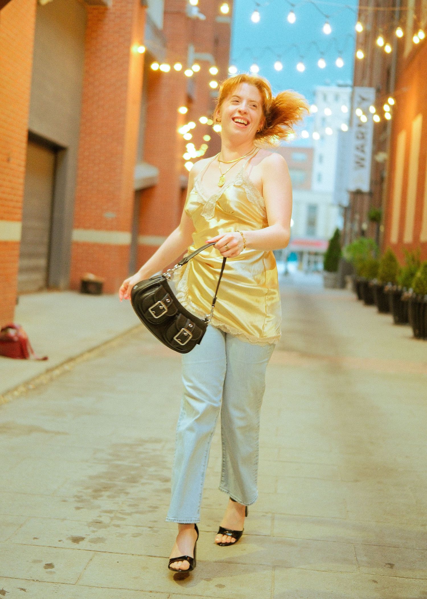 A smiling woman in a gold top and light jeans walks down a city alley with string lights, carrying a black bag.