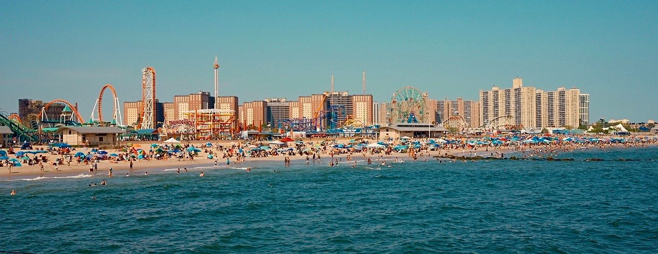 Panoramic view of Coney Island beach with crowds, amusement park rides, and city skyline on a sunny day.