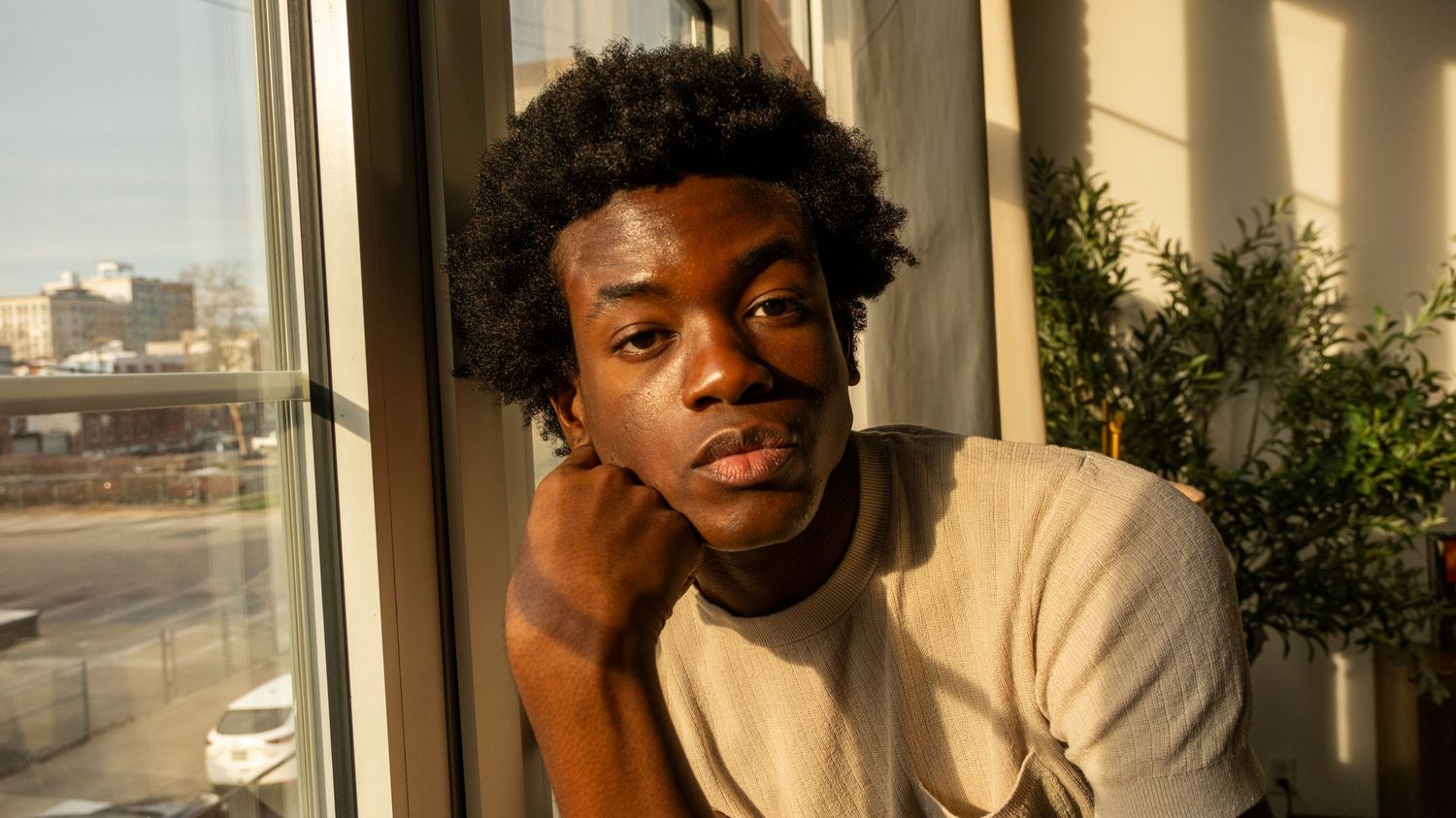 Young Black man with curly hair leaning on hand by a sunny window in a beige shirt.