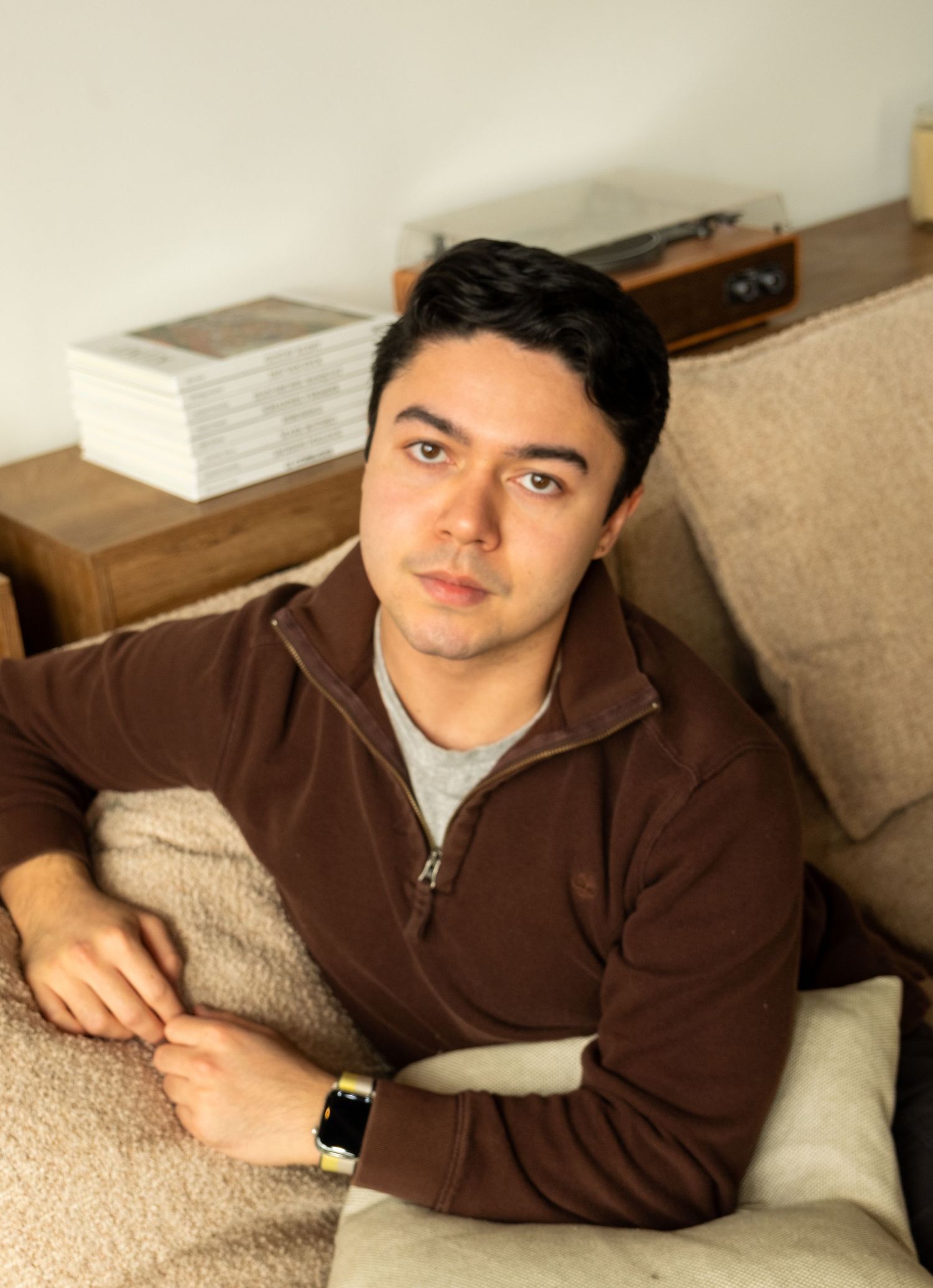 Young man in brown zip-up hoodie sitting relaxed on a couch wearing a gold watch.
