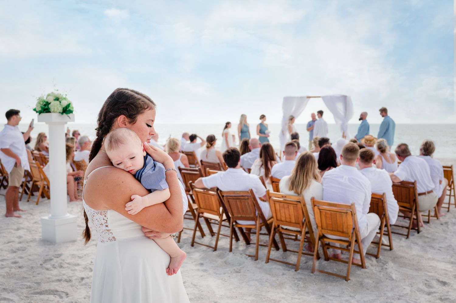 A beach wedding ceremony with guests seated in wooden chairs on white sand near the ocean.