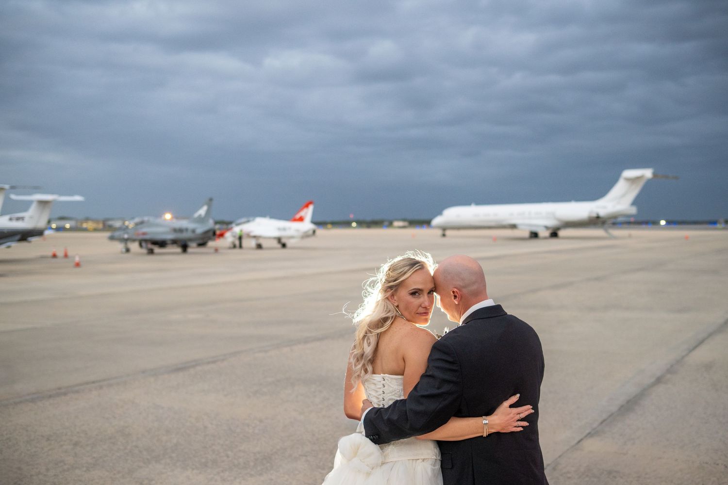 Bride and groom embrace on airport tarmac with jets and stormy sky backdrop on their wedding day.