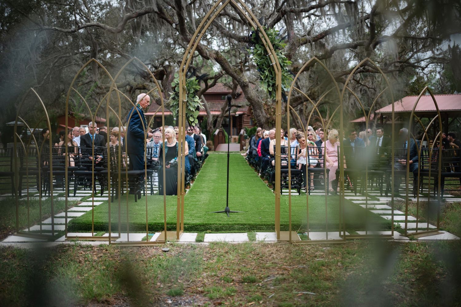 Outdoor wedding ceremony with gold arch decor, green aisle, and guests seated among moss-draped trees.