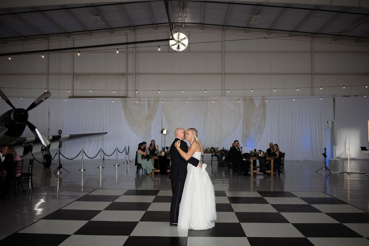 Bride and groom share first dance on checkered floor in airplane hangar wedding venue.