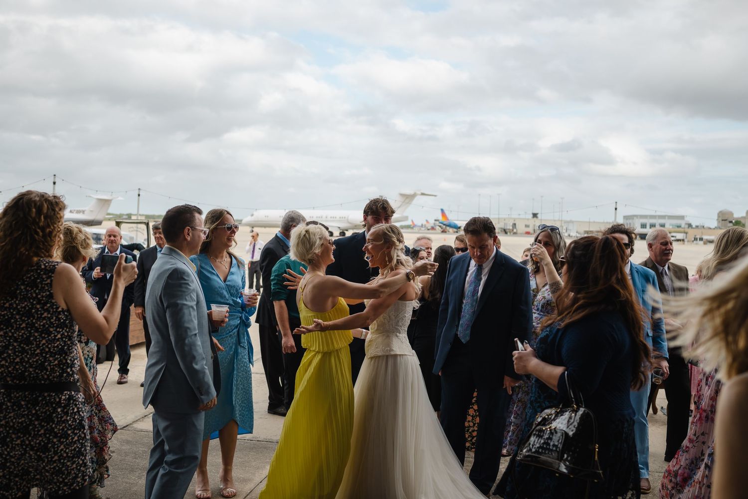 Wedding guests celebrate outdoors on an airport tarmac, with a bride in white dancing among elegantly dressed attendees.