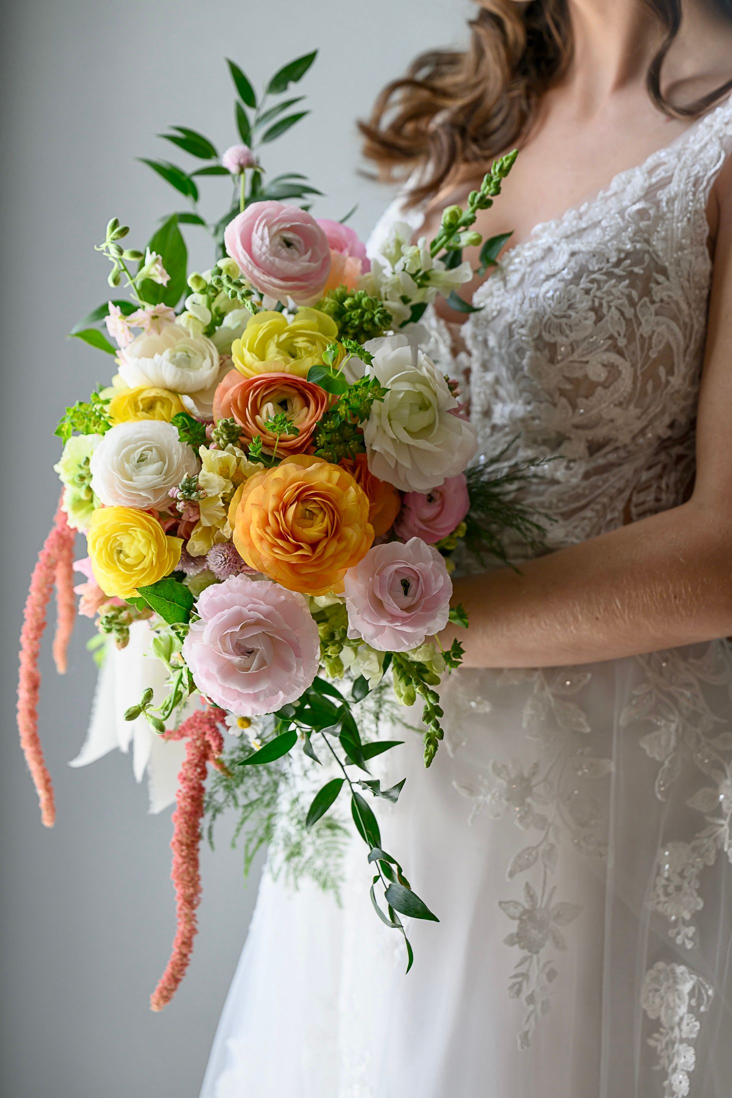 A romantic bridal bouquet featuring pink, yellow and white ranunculus flowers with green foliage against a lace wedding dress.