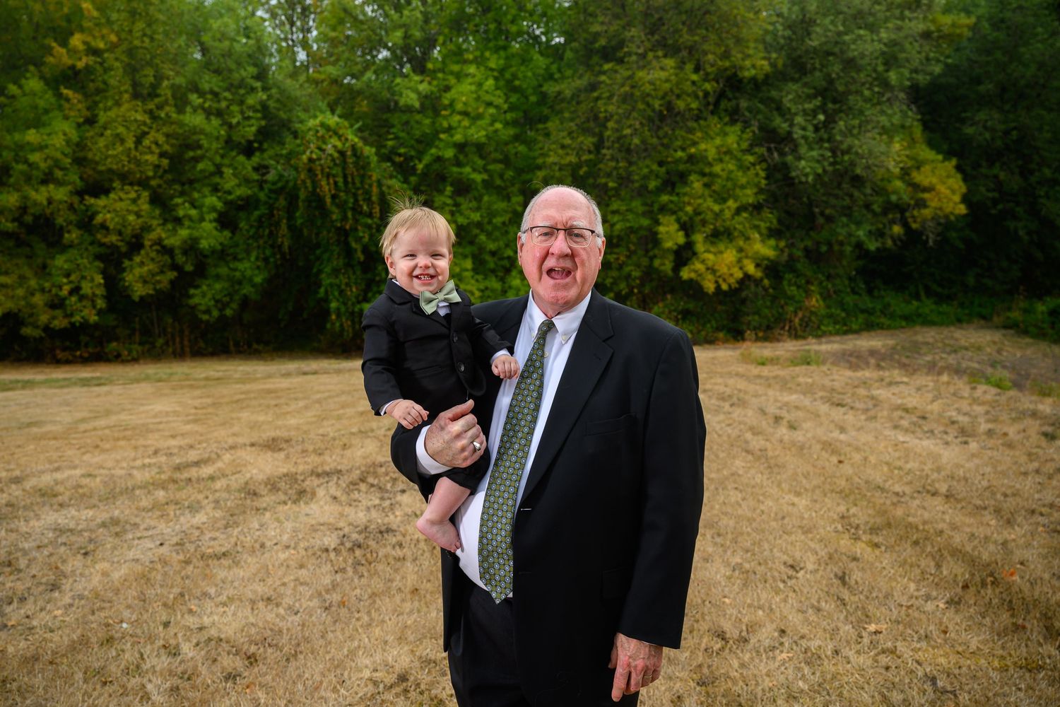 Two people in formal attire stand together in an outdoor setting with autumn trees in the background.