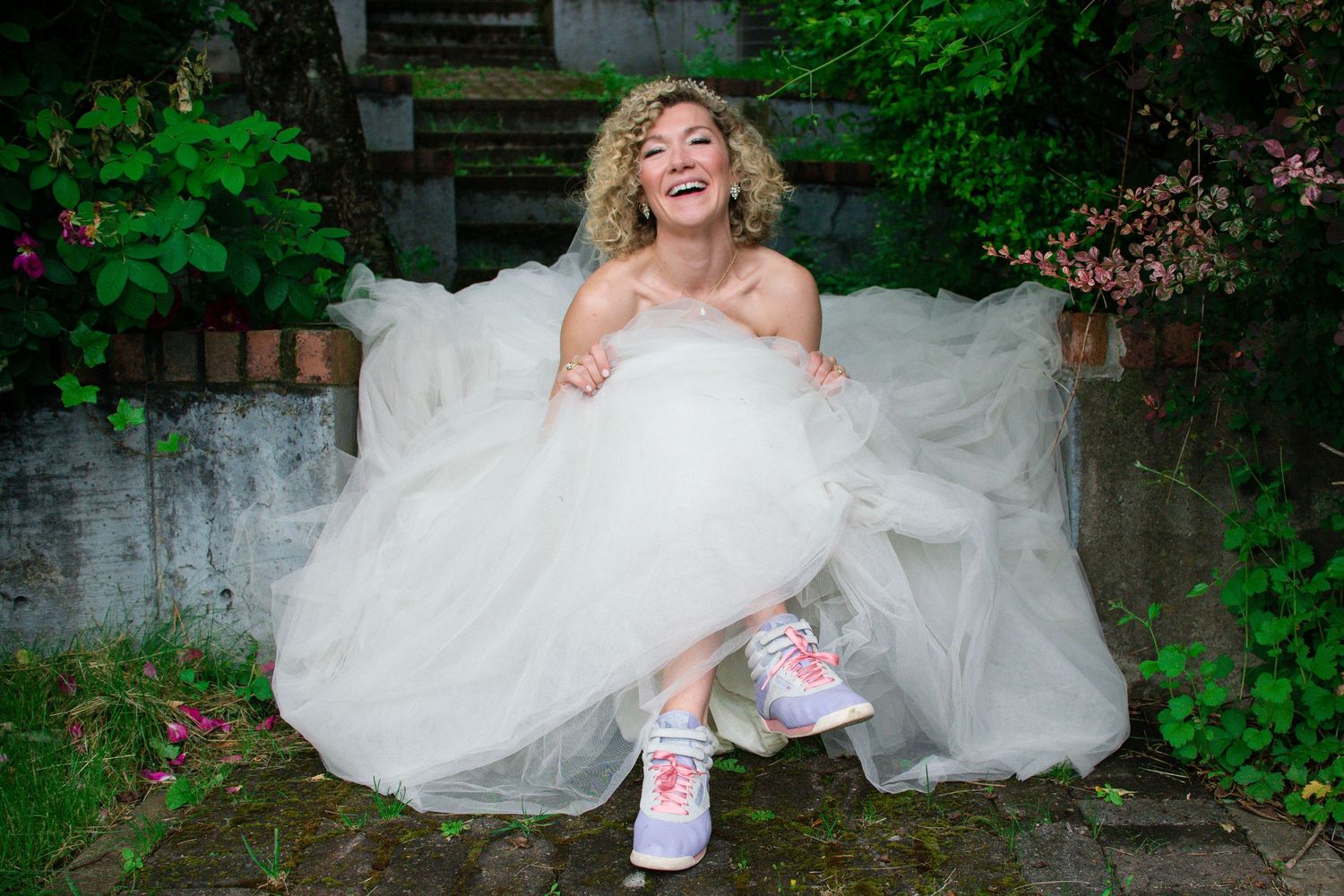 Bride in white dress sitting playfully in a lush garden setting.