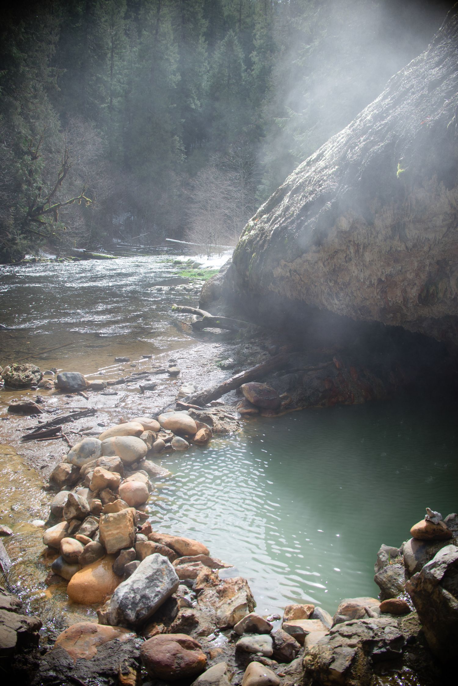 Steamy natural hot spring pool surrounded by rocks and boulders in a misty forest setting.