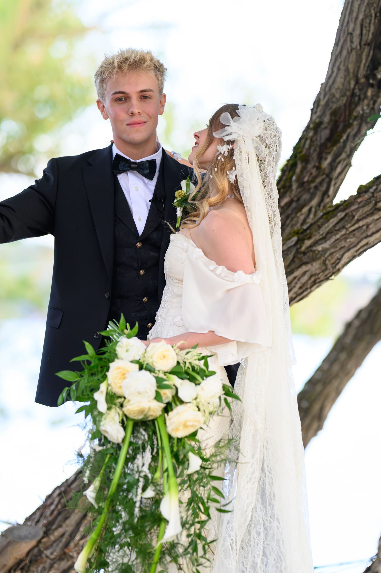Bride in lace veil holding white peony bouquet poses with groom in black tuxedo beside a tree outdoors.