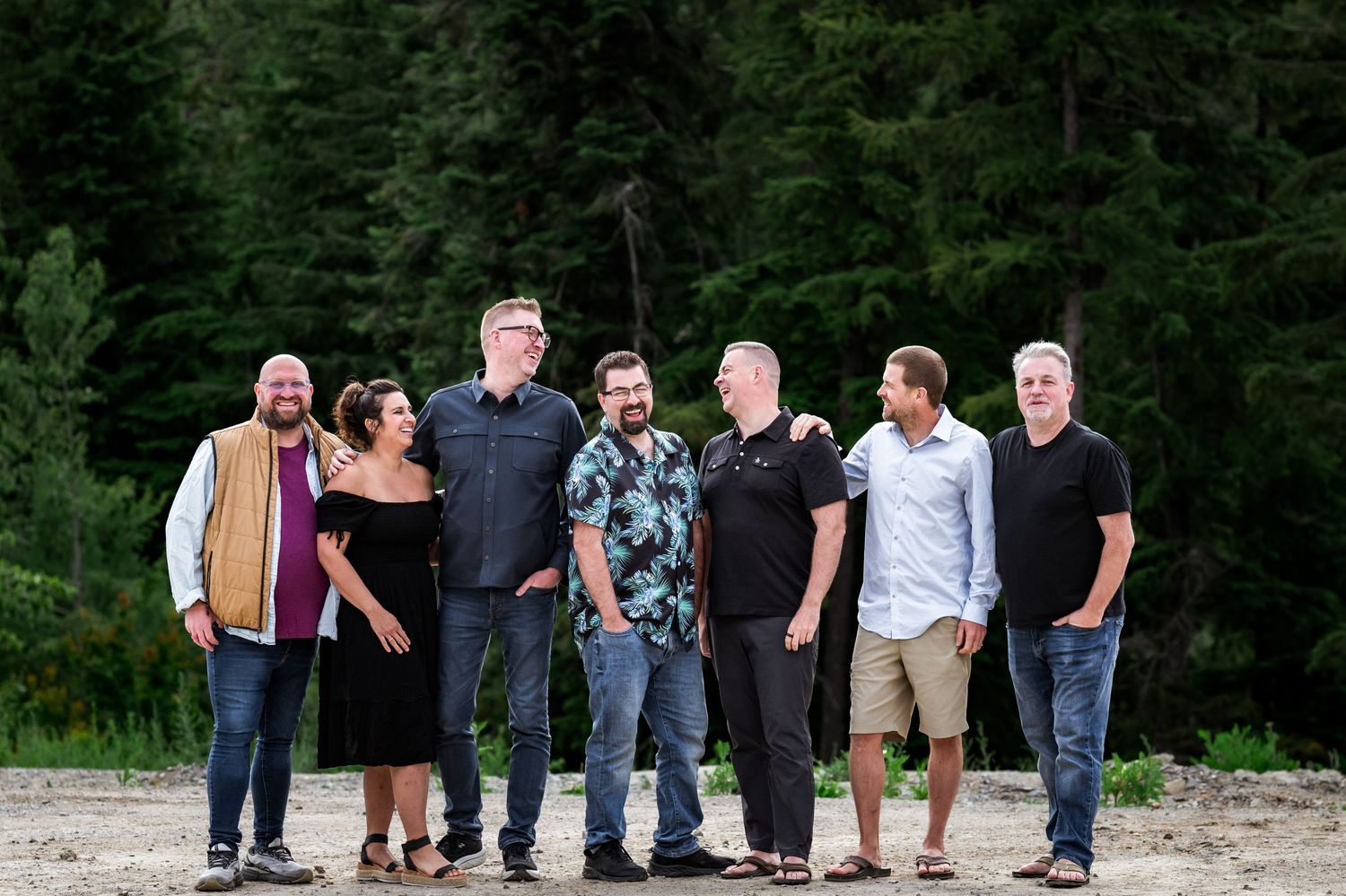 Seven adults smiling and posing together outdoors with tall green pine trees in the background on a sunny day.