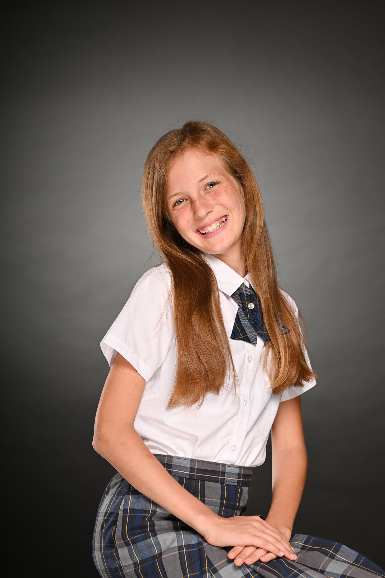 Smiling young girl in school uniform with white shirt, plaid skirt, and navy bow tie poses against gray background.