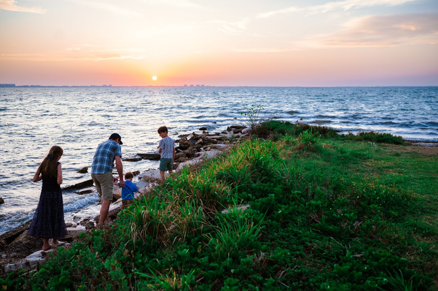 People enjoying sunset by the ocean on a grassy cliff with rocky shoreline during golden hour.