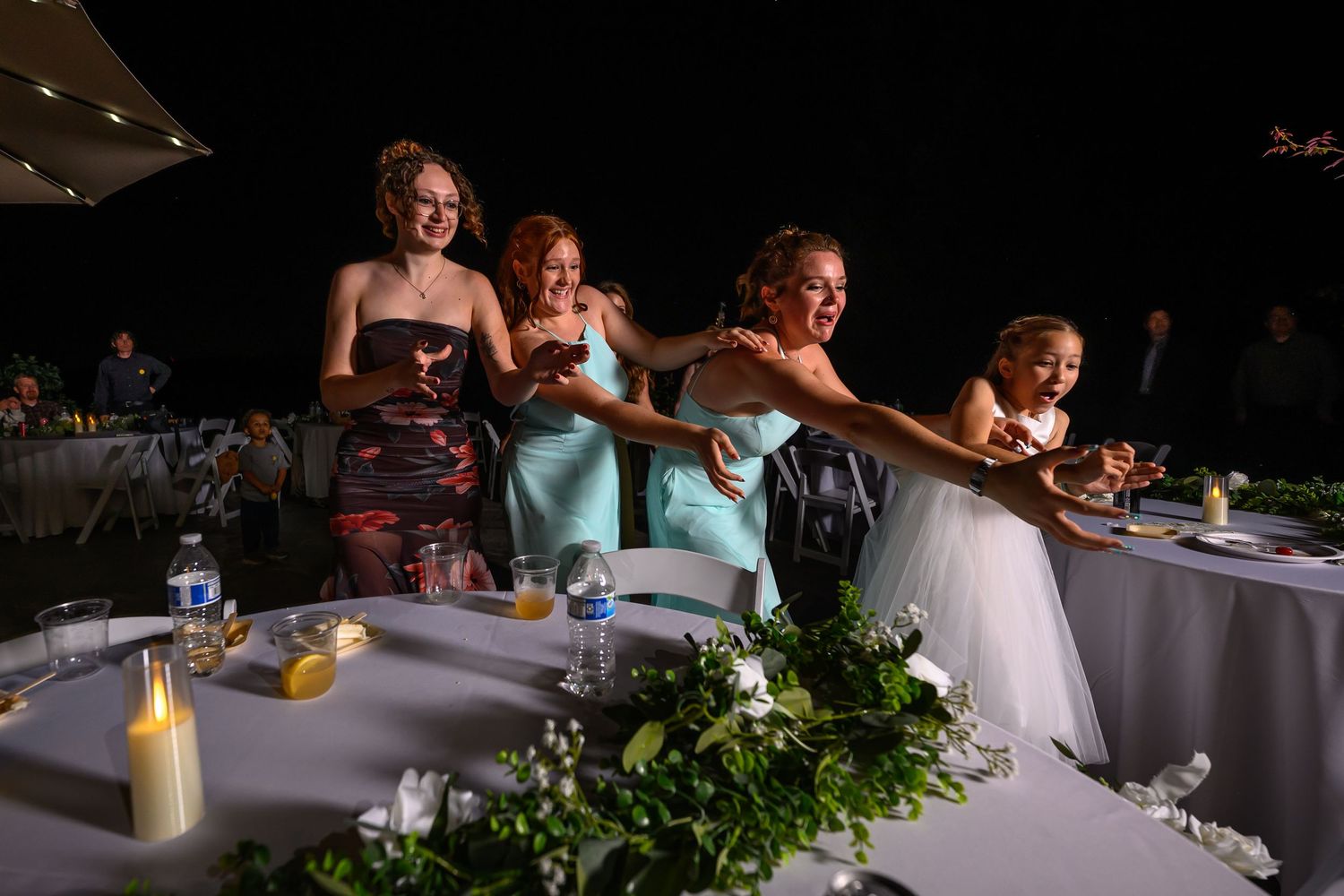 Wedding guests laugh and socialize at a reception table decorated with floral centerpieces and white linens.