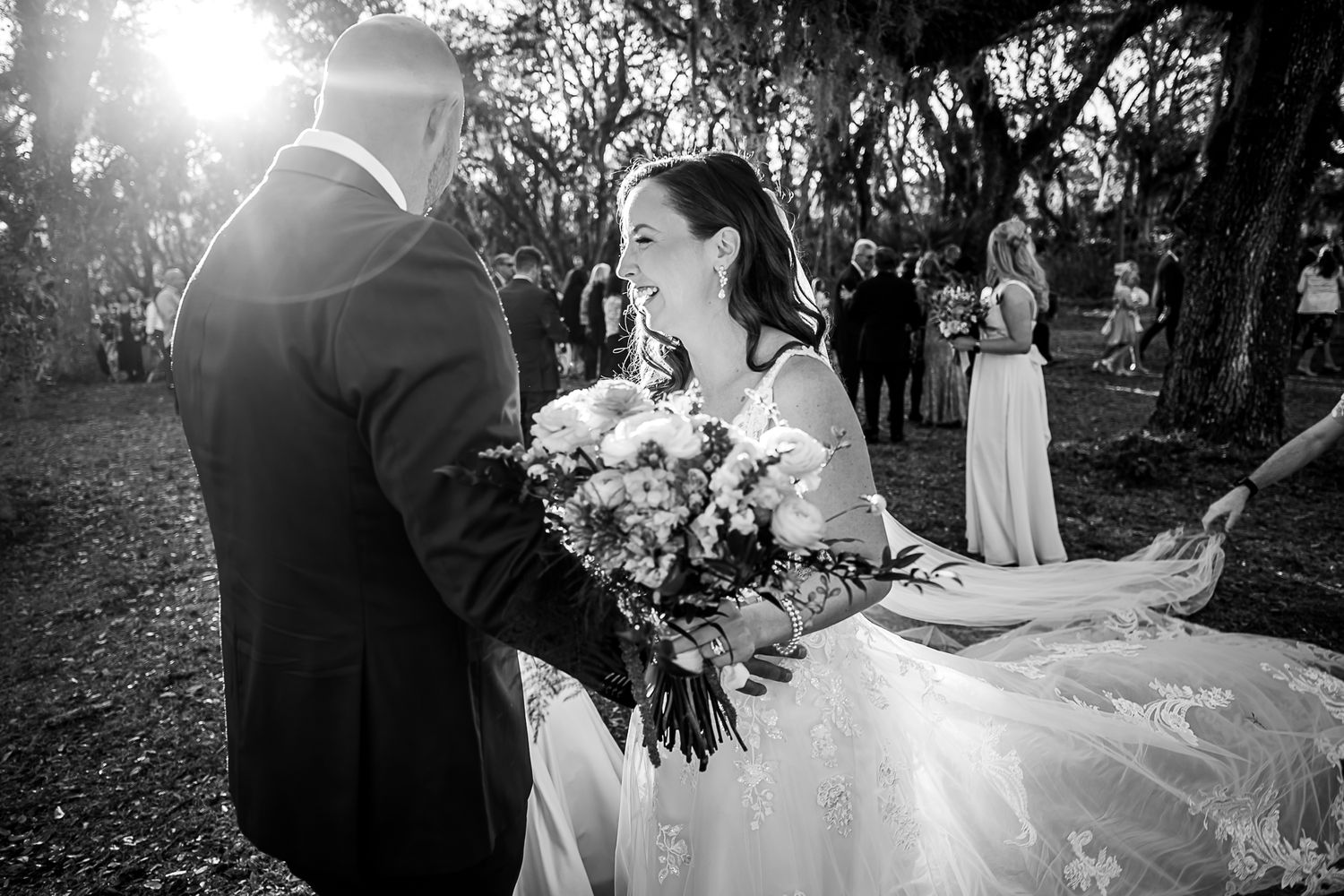 A bride in a white gown holds a floral bouquet as sunlight filters through trees during an outdoor wedding ceremony.