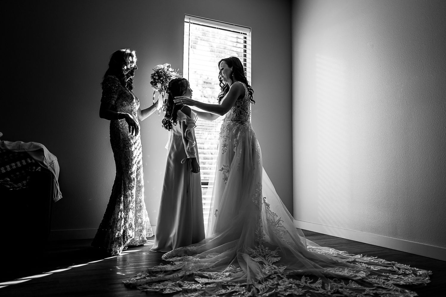 Three women in elegant wedding gowns stand together by a window in a dramatic black and white photograph.