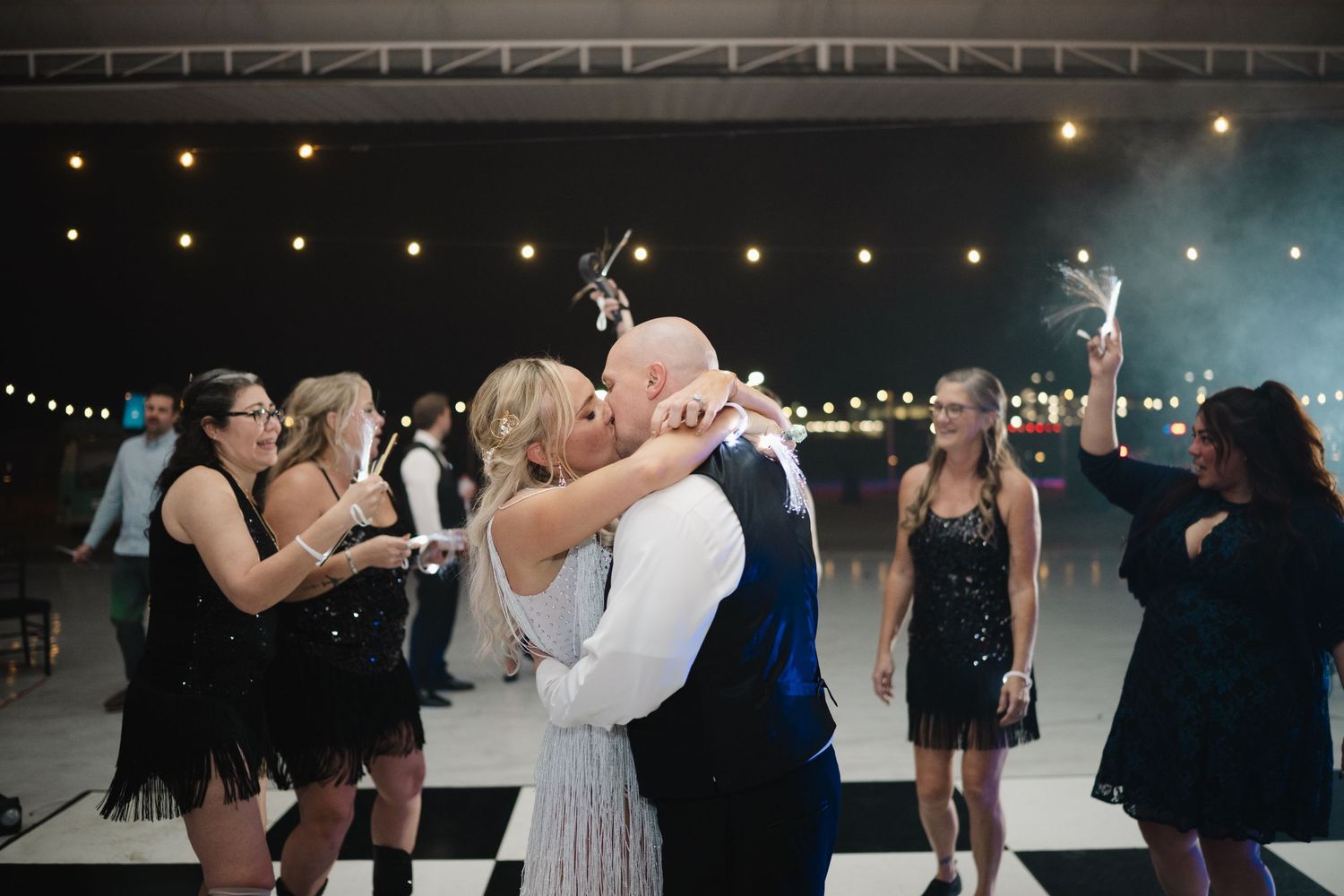Bride and groom share a kiss on dance floor surrounded by bridesmaids holding sparklers at outdoor wedding reception.