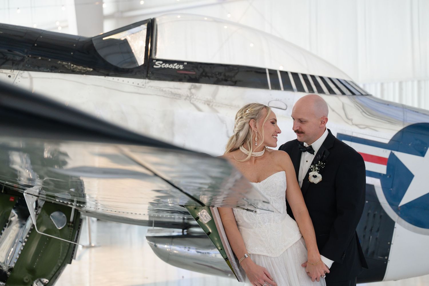 Bride and groom share a tender moment next to a classic military aircraft in a hangar.