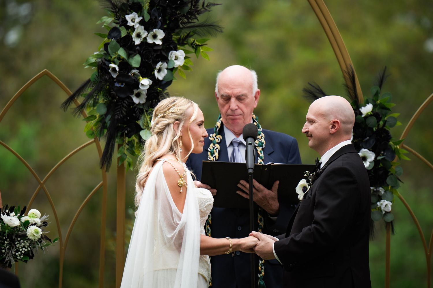 Bride and groom exchange vows at outdoor wedding ceremony beneath floral arch with officiant.