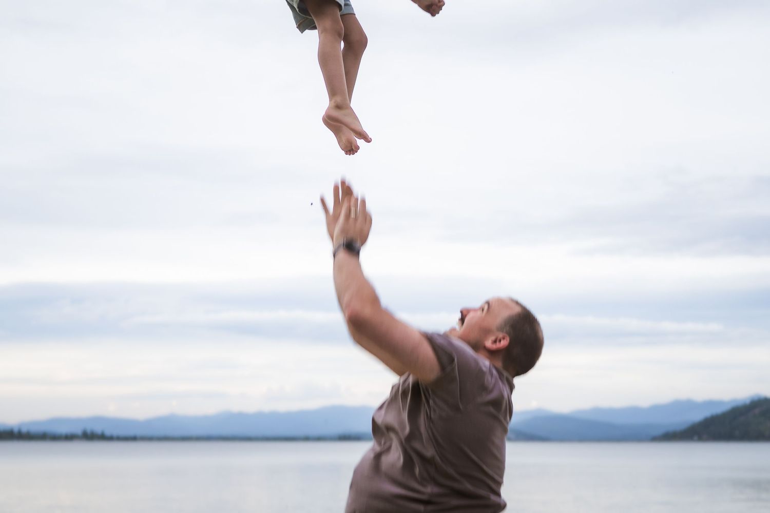 A father joyfully tosses his child into the air near a serene lake with mountain views in the background.