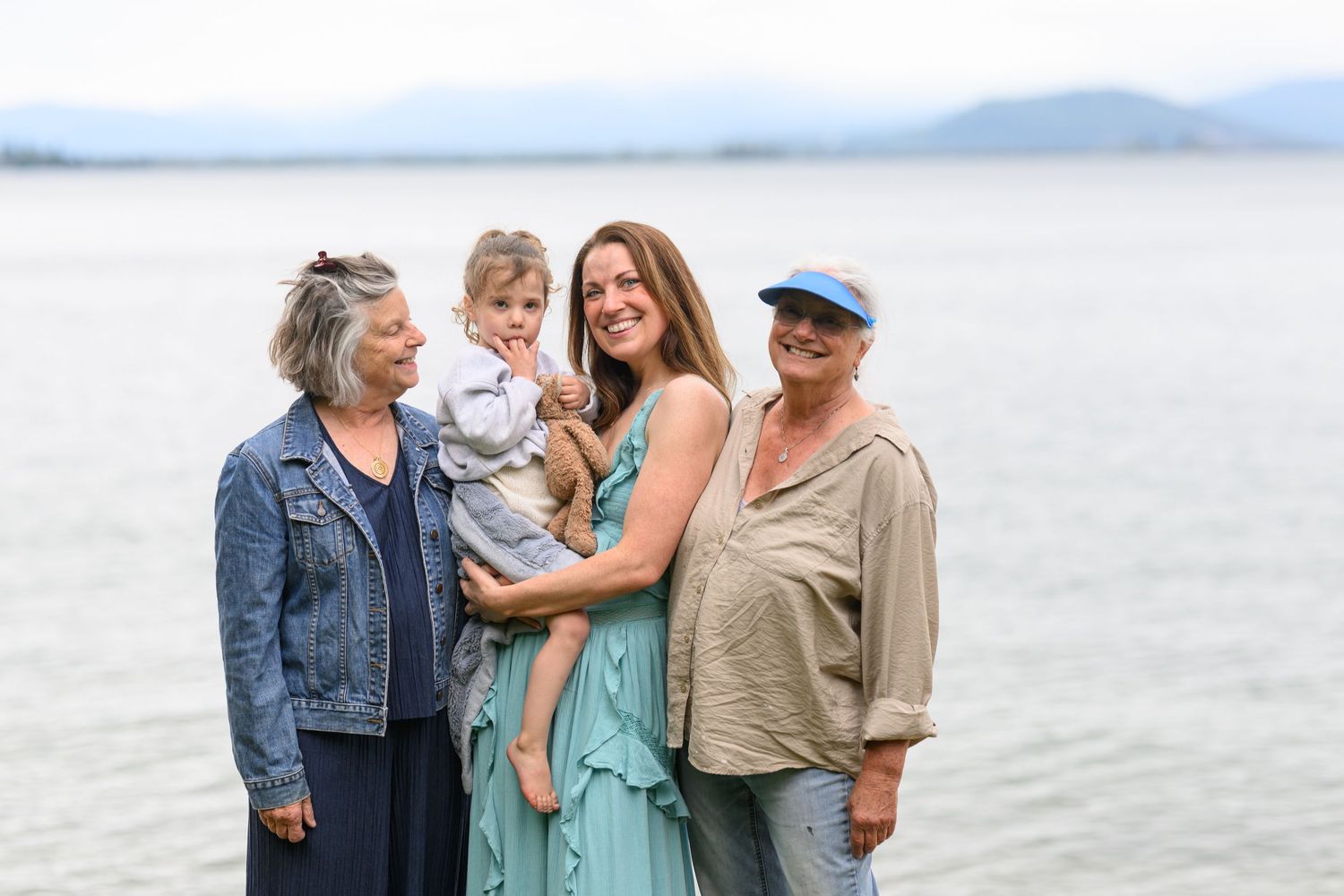 Multi-generational family of four posing together outdoors by a lake, smiling with mountains in the background.