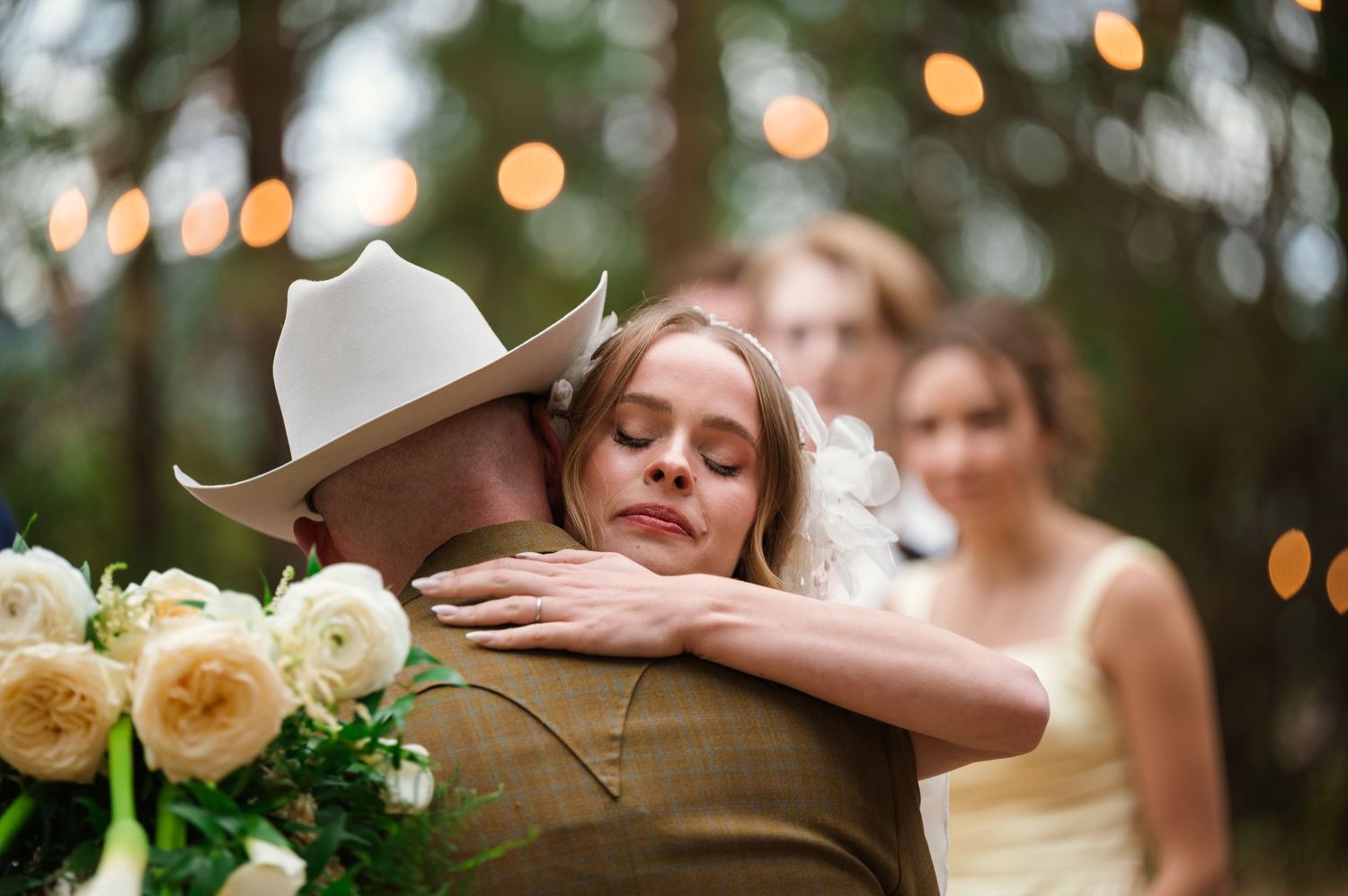Bride hugging groom in cowboy hat at outdoor wedding ceremony with bokeh lights and bridesmaids in background.