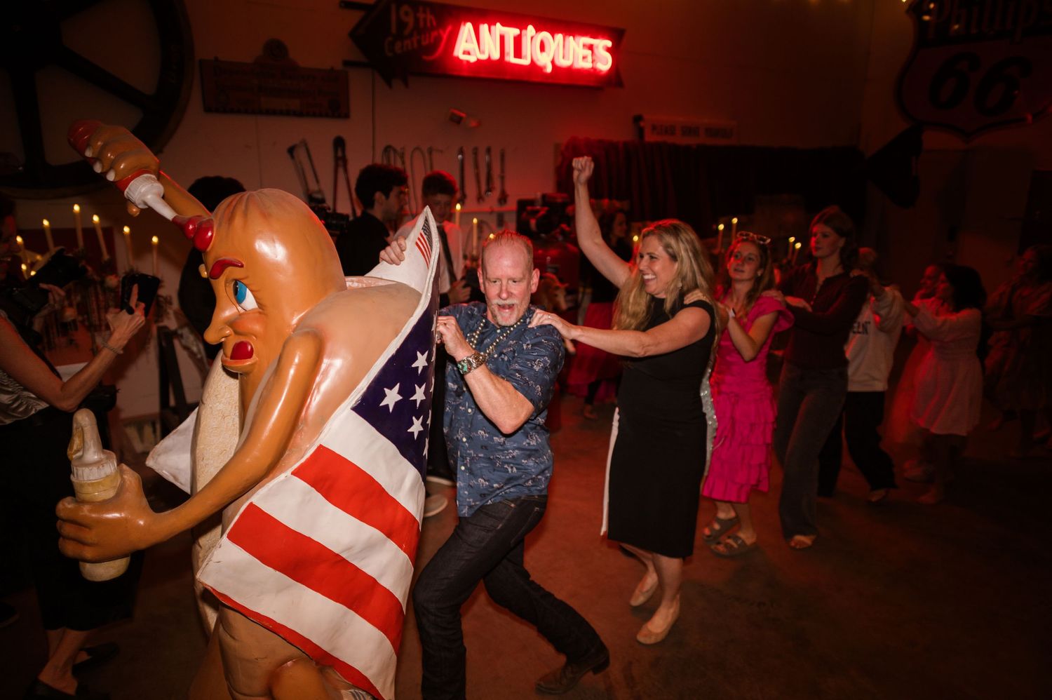 People dancing at a lively party near an Antiques sign, with someone in a patriotic hot dog costume.