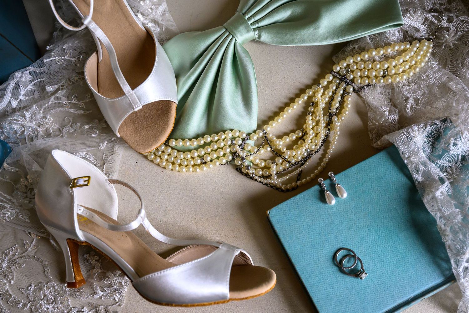 Elegant bridal accessories including pearl necklaces, satin heels, and delicate lace details arranged on a light surface.