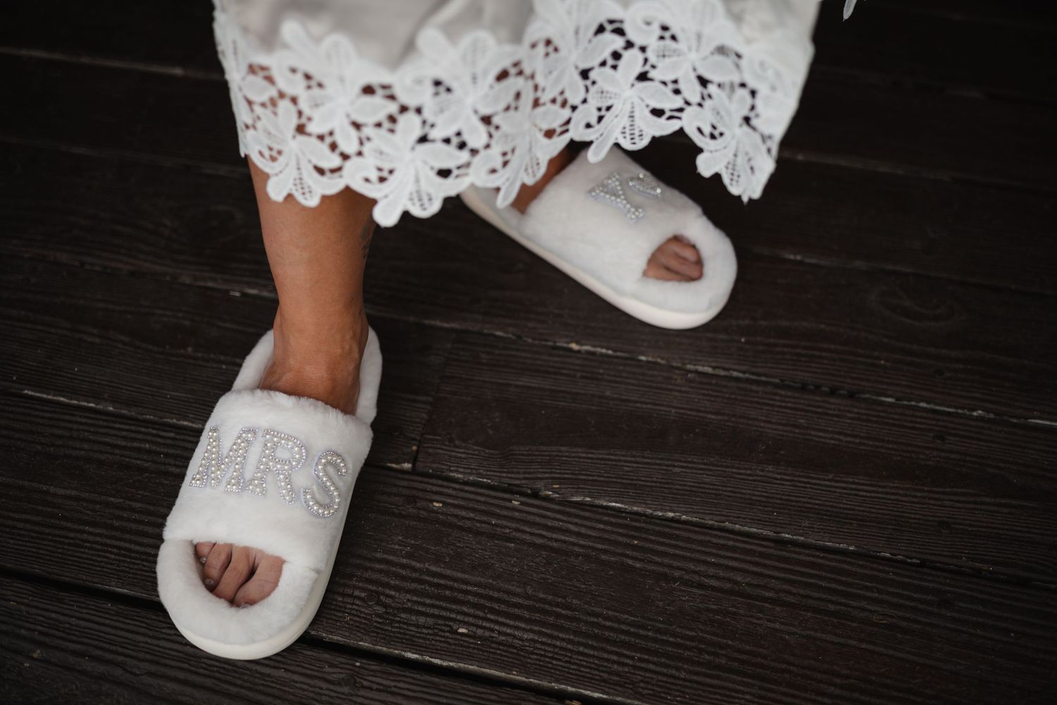 Bride wearing white fluffy MRS slippers with rhinestone letters on dark wood floor with lace dress hem.