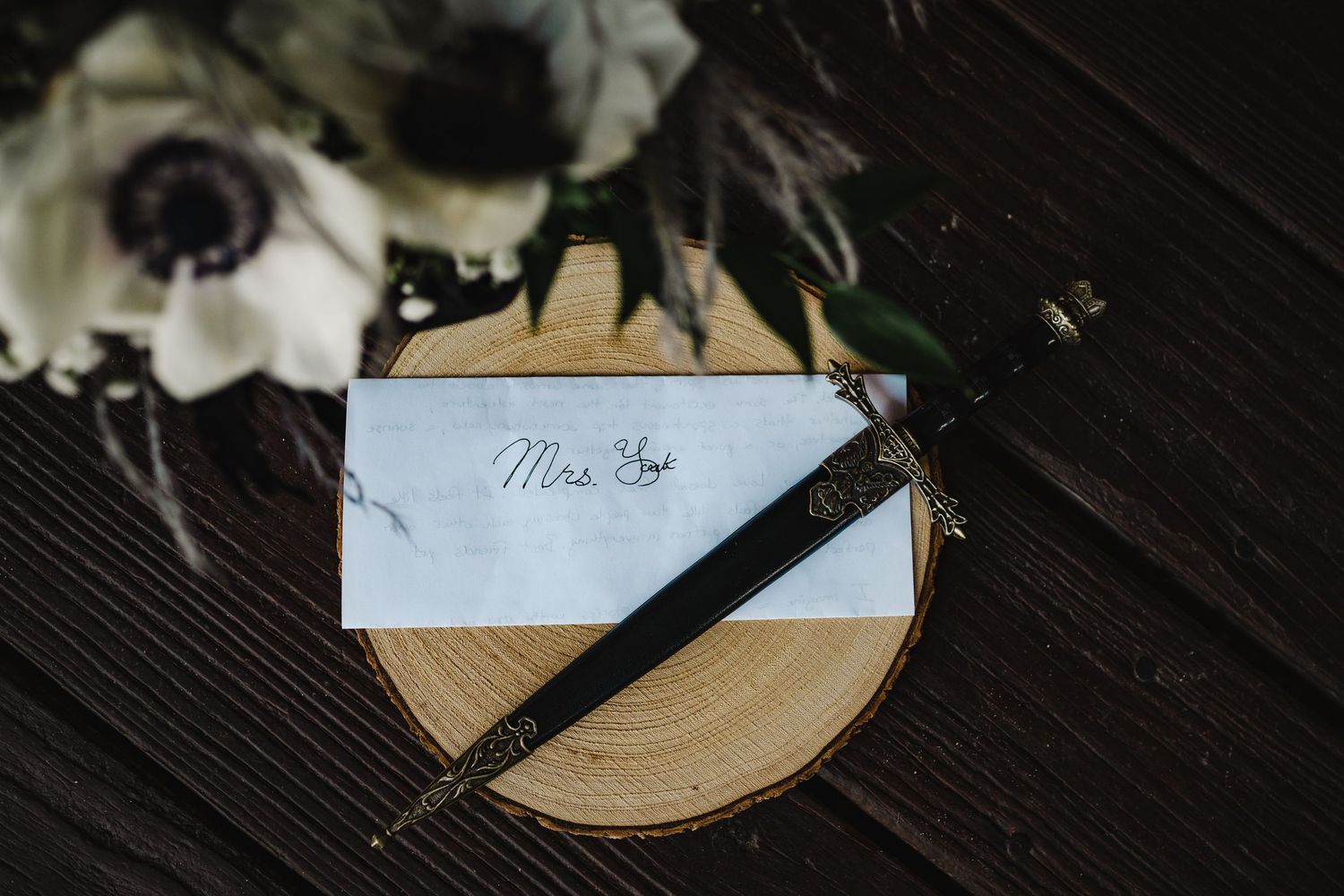 A calligraphy Mrs. card with a pen on a wood slice next to white anemone flowers at a wedding.