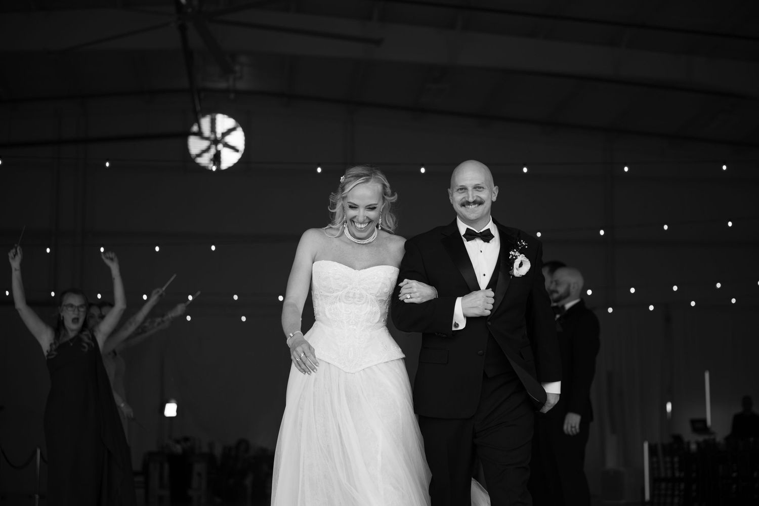 Black and white photo of a smiling bride in strapless gown and groom in tuxedo entering their wedding reception.