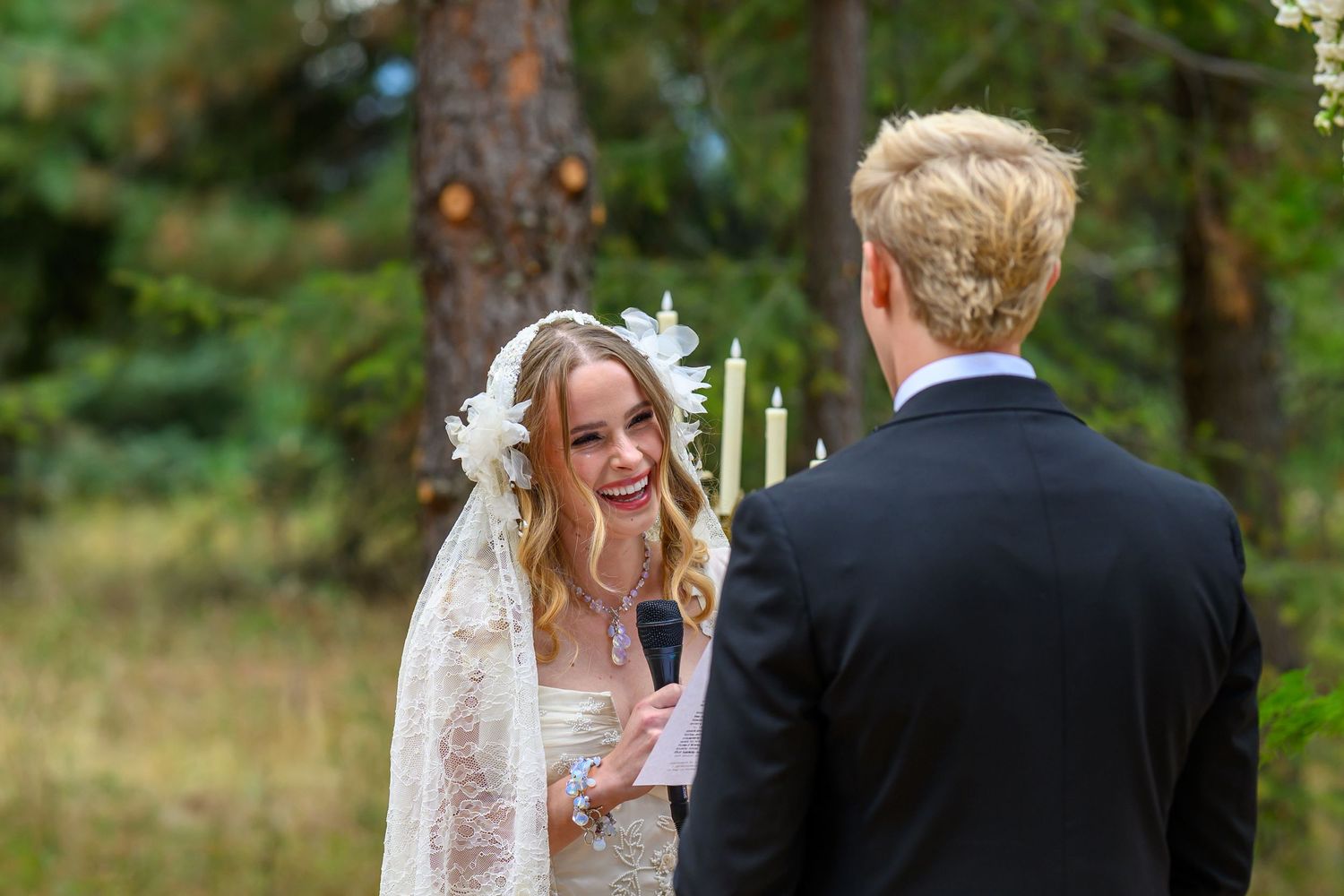 Bride in lace veil smiles at groom during outdoor wedding ceremony surrounded by pine trees.