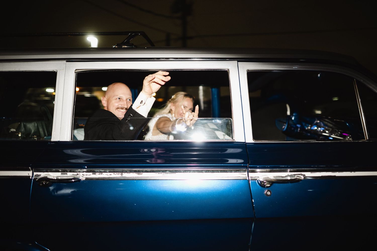 Couple waving from a vintage blue classic car at night, celebrating a wedding getaway.