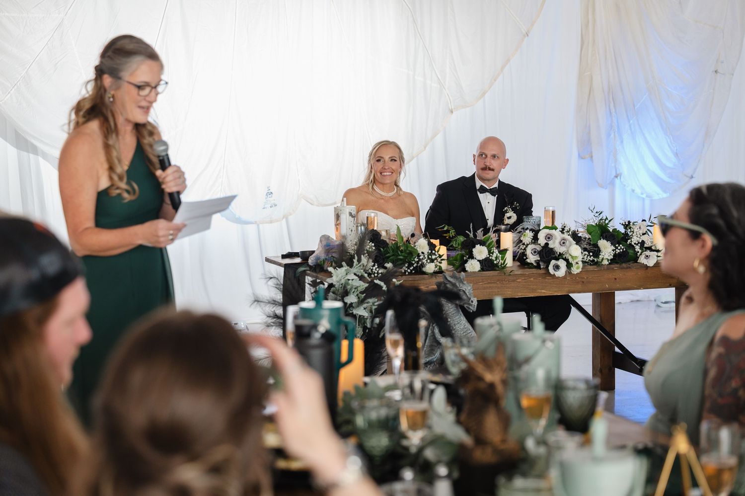 Bridesmaid in green dress gives speech at wedding reception while couple sits at floral sweetheart table.