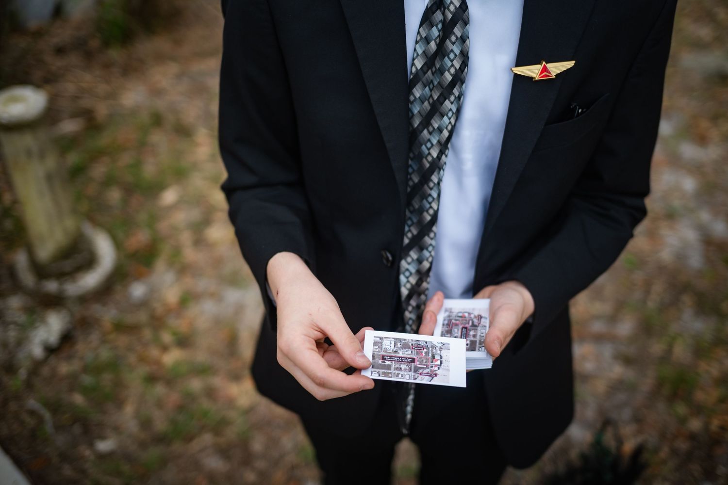 Groom in black suit holding small photo cards at outdoor wedding venue.