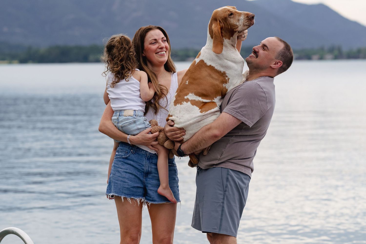 Happy family with young child and brown white dog posing by mountain lake on cloudy day.