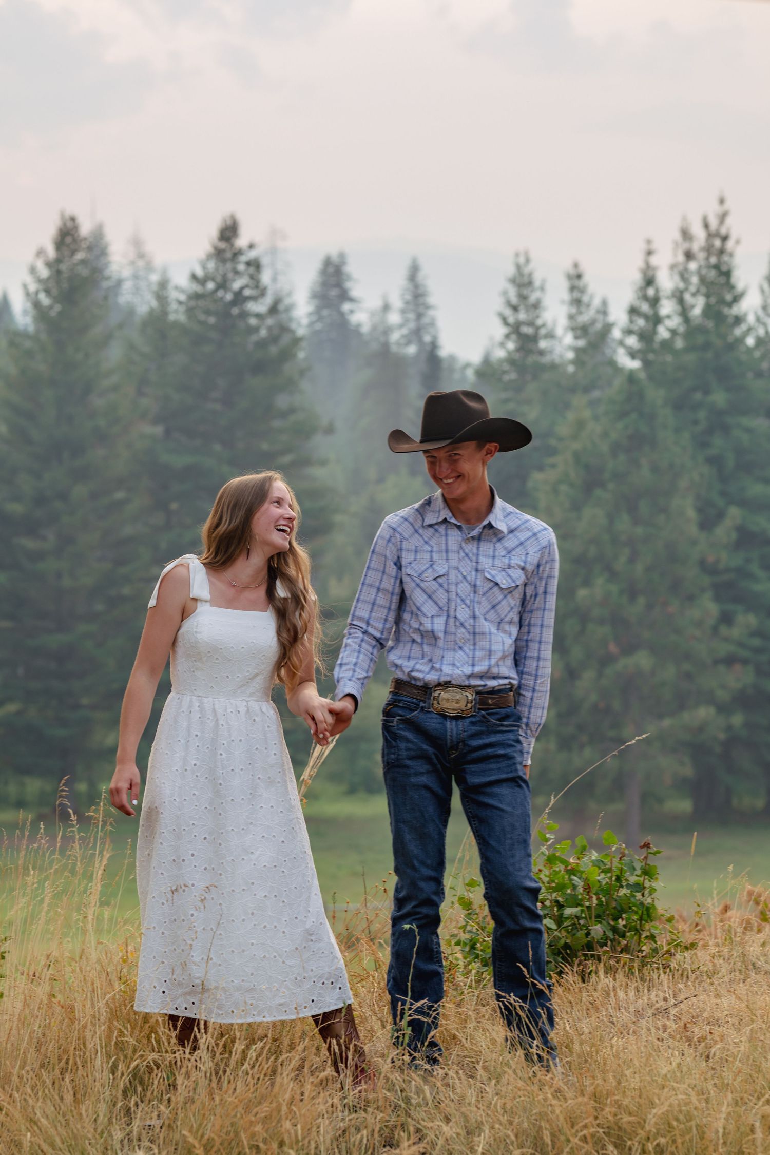 A young couple in western attire holds hands in a golden field, smiling, with pine trees in the background.