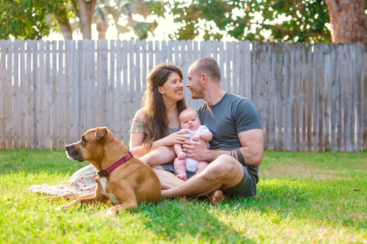 Family relaxing on grass with their golden retriever in a fenced backyard.