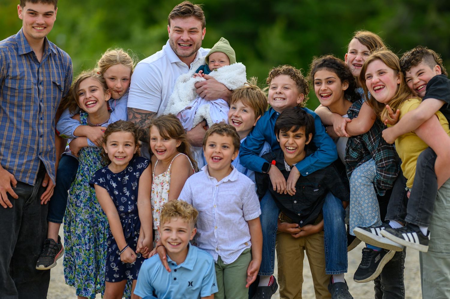 Large family group photo with man holding baby surrounded by many smiling children outdoors.