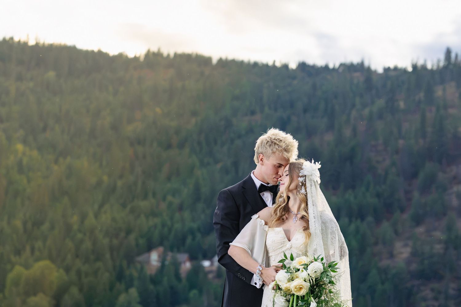 Bride and groom share a romantic kiss on a mountain overlook surrounded by lush pine forests on their wedding day.
