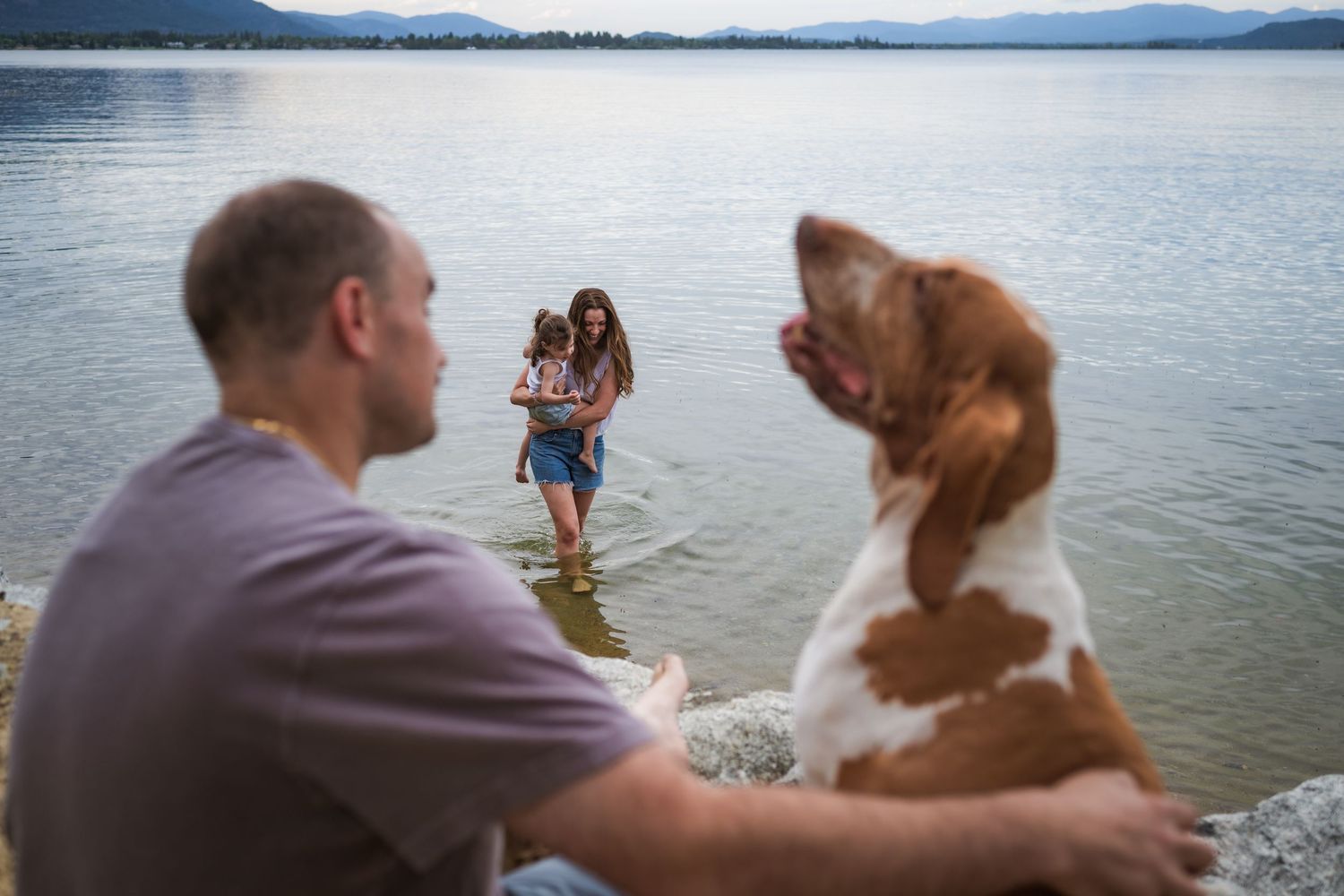 A family enjoys a lakeside outing with their brown and white dog in the foreground as a woman and child wade in calm water.
