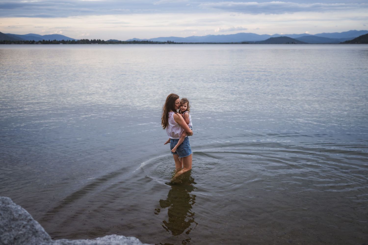 Two girls standing in a lake at dusk, embracing near the shoreline with mountains in the background.