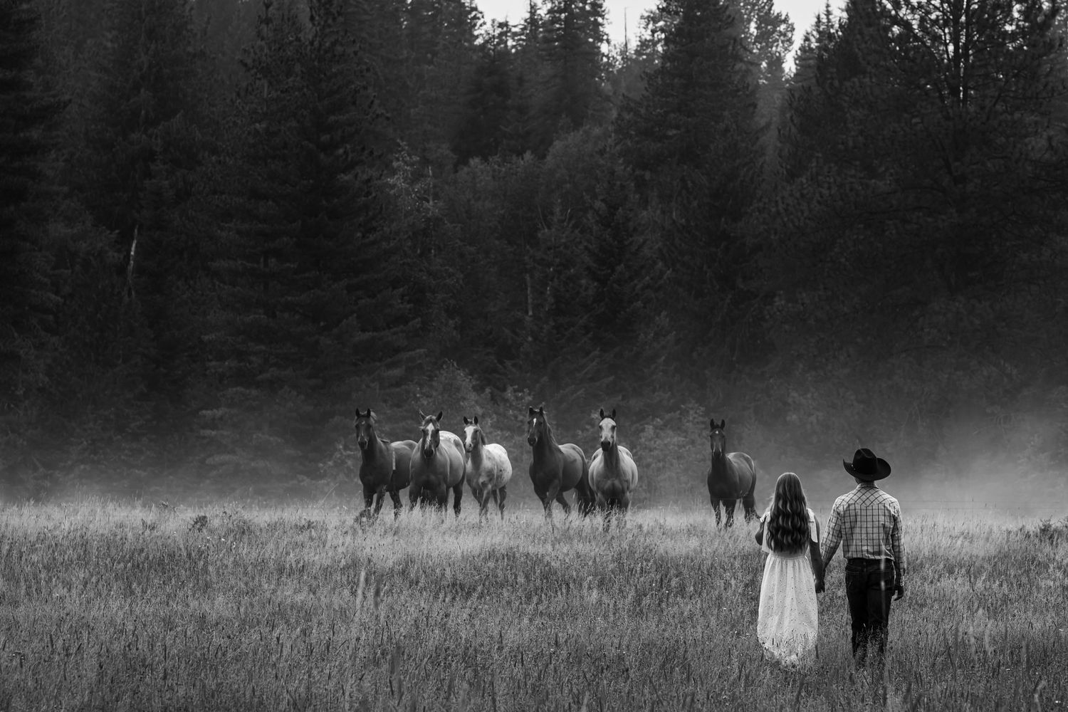 Black and white photo of a couple holding hands watching horses run through a misty field near a forest.