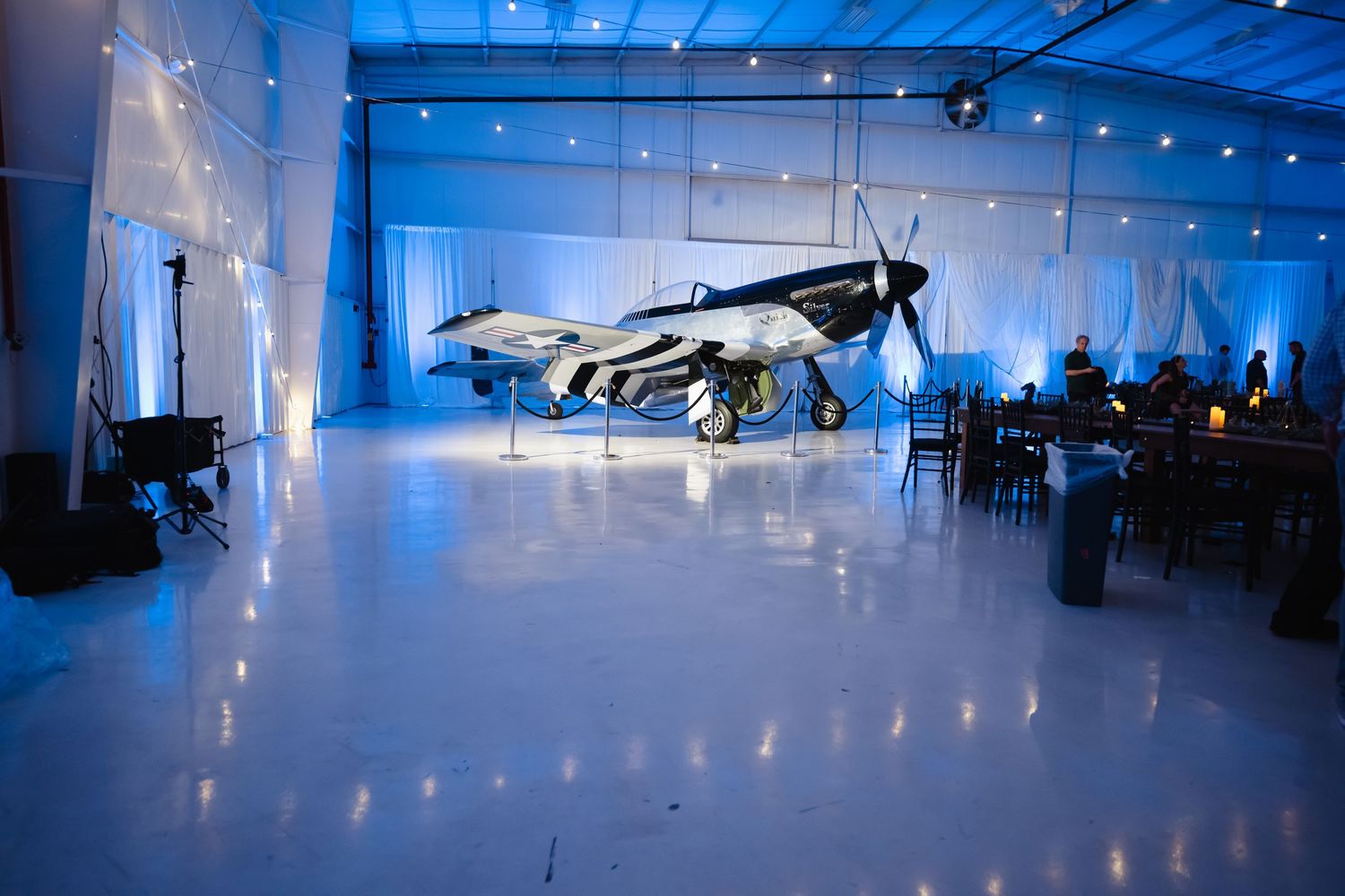 Vintage WWII fighter plane displayed as wedding venue centerpiece in blue-lit hangar with elegant reception tables.