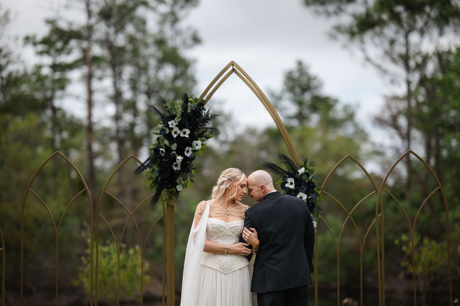 Bride and groom share first kiss under gold geometric arch with black and white floral arrangements in a forest wedding ceremony.