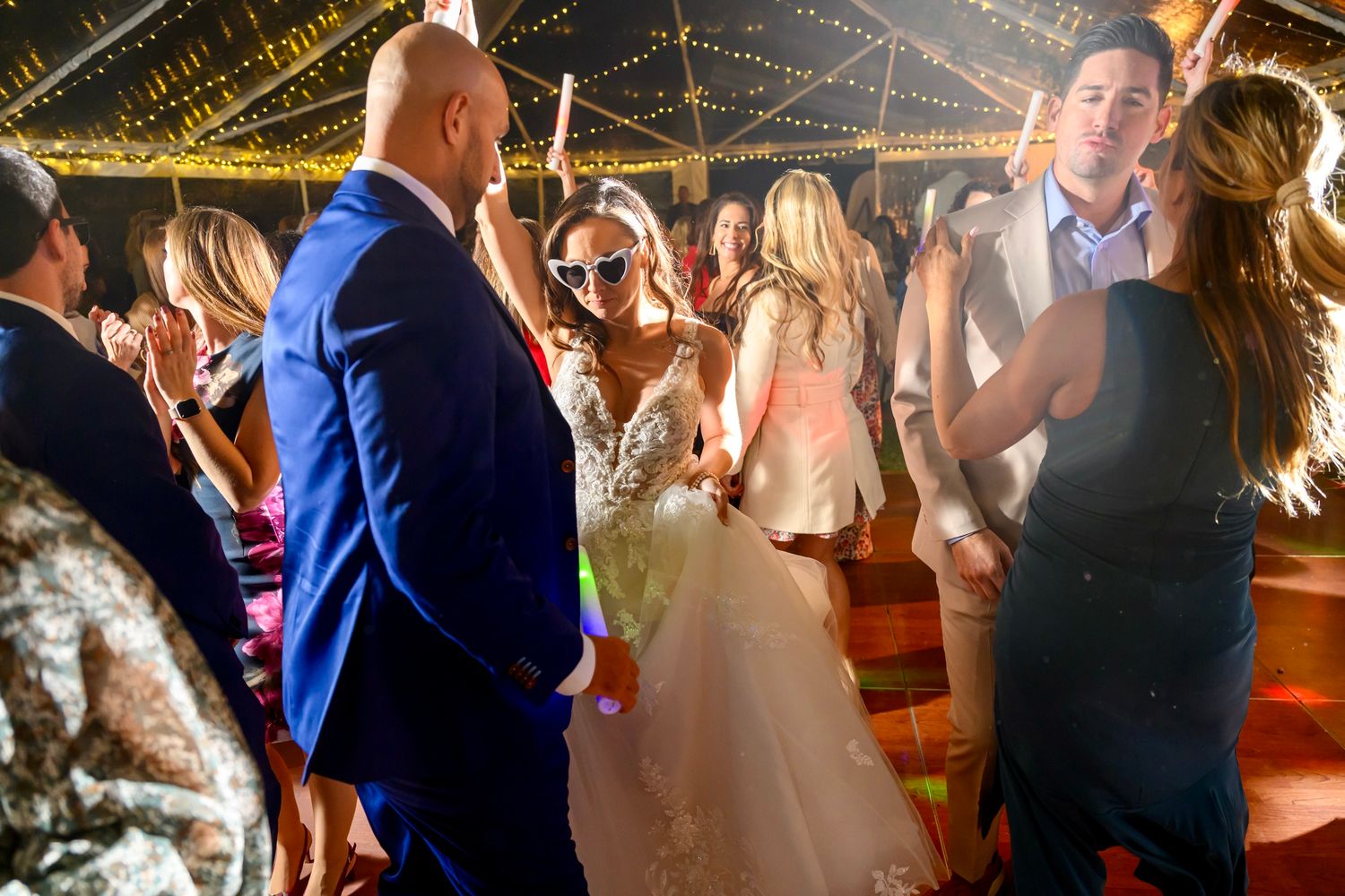 Wedding guests and newlyweds dancing together under twinkling lights at a festive evening reception celebration.