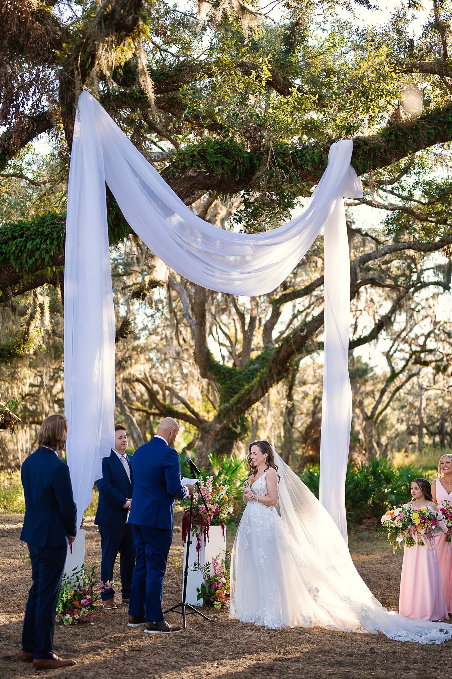 Outdoor wedding ceremony under oak trees with white fabric draped arch and floral arrangements in a natural setting.